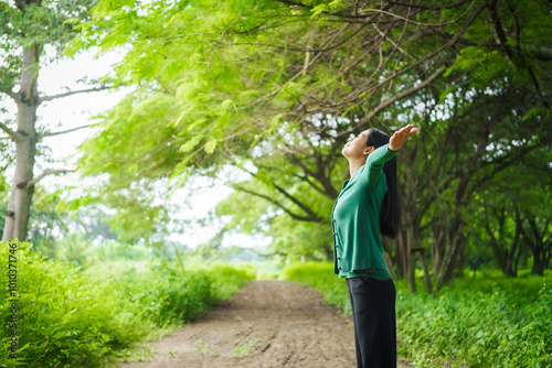 An Asian woman is standing happily in a green forest park, enjoying the fresh air and nature. She takes a deep breath, looks up at the morning sunlight, and feels relaxed and peaceful.