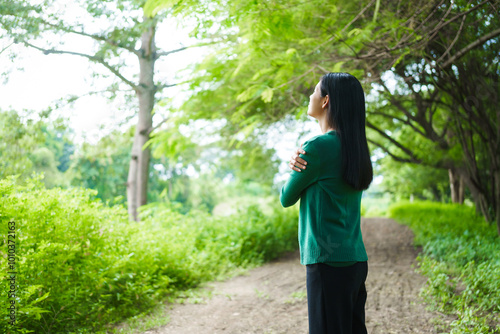 An Asian woman is standing happily in a green forest park, enjoying the fresh air and nature. She takes a deep breath, looks up at the morning sunlight, and feels relaxed and peaceful.