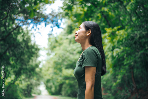 An Asian woman is standing happily in a green forest park, enjoying the fresh air and nature. She takes a deep breath, looks up at the morning sunlight, and feels relaxed and peaceful.