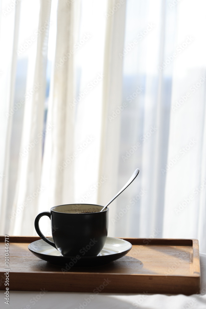 Fototapeta premium A black mug filled with steaming coffee sits on a wooden table, illuminated by the sun streaming in through a nearby window.