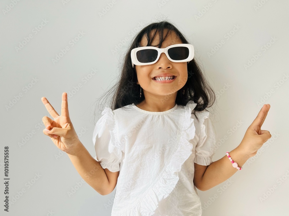 Five year old Asian child wearing white shirt raising hands making peace gesture using white glasses Five year old Asian child wearing white shirt raising hands making peace gesture using white glasses