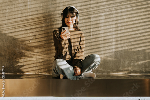 Sunlit Moment: A young woman, radiating a sense of peace, sits cross-legged against a wall, bathed in warm sunlight streaming through blinds. She holds a phone and wears headphones.