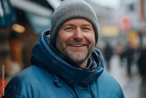 Man wearing a blue jacket and a gray hat is smiling. He is standing in the rain