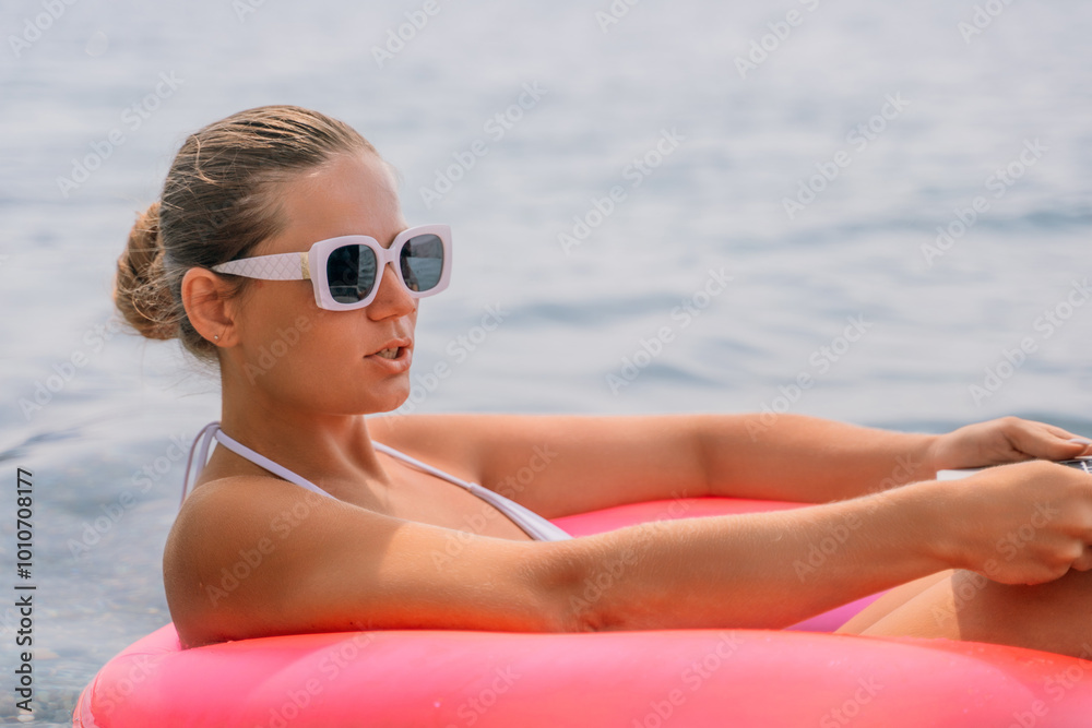 A woman is sitting in a pink inflatable raft on a lake, holding a laptop. Concept of relaxation and leisure, as the woman enjoys her time in the water while working on her laptop. A woman is sitting in a pink inflatable raft on a lake, holding a laptop. Concept of relaxation and leisure, as the woman enjoys her time in the water while working on her laptop.