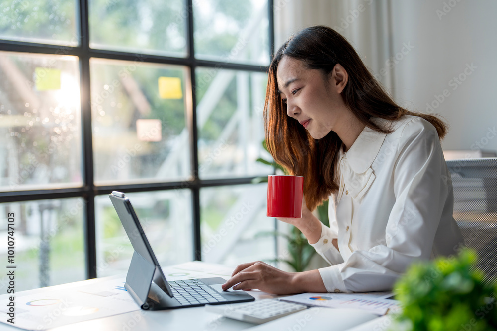 Young asian businesswoman drinking coffee and working on tablet in modern office Young asian businesswoman drinking coffee and working on tablet in modern office
