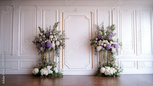 A white wall with classic wainscoting is decorated with two tall arrangements of purple and white flowers  on gold stands.
