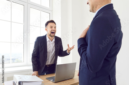 Young angry businessman wearing suit shouting on a stressed man company employee sitting at desk on workplace with laptop in office. Angry boss yelling at his male worker. Business conflict concept.