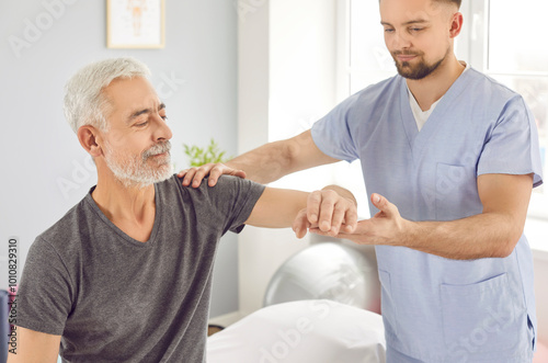 Young male physiotherapist helps old white haired retired man regain elbow mobility after stroke. Physiotherapy clinic, professional rehabilitation for senior patients concept photo