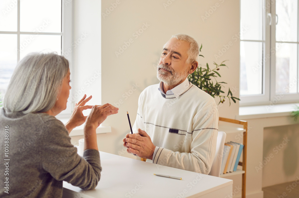 Gary-haired elderly couple talking sitting at table in living room and discussing something indoors. Senior man and woman communicating at home. Family relationship in retirement concept Gary-haired elderly couple talking sitting at table in living room and discussing something indoors. Senior man and woman communicating at home. Family relationship in retirement concept