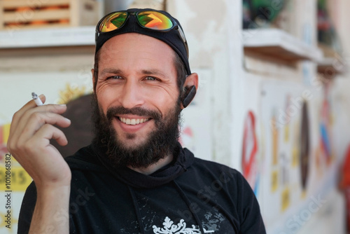 Handsome 45 years man with a prominent beard and sunglasses on their head sits comfortably outdoors, holding a cigarette in his hand. Dressed in a dark hoodie and sporting a relaxed expression, Israel