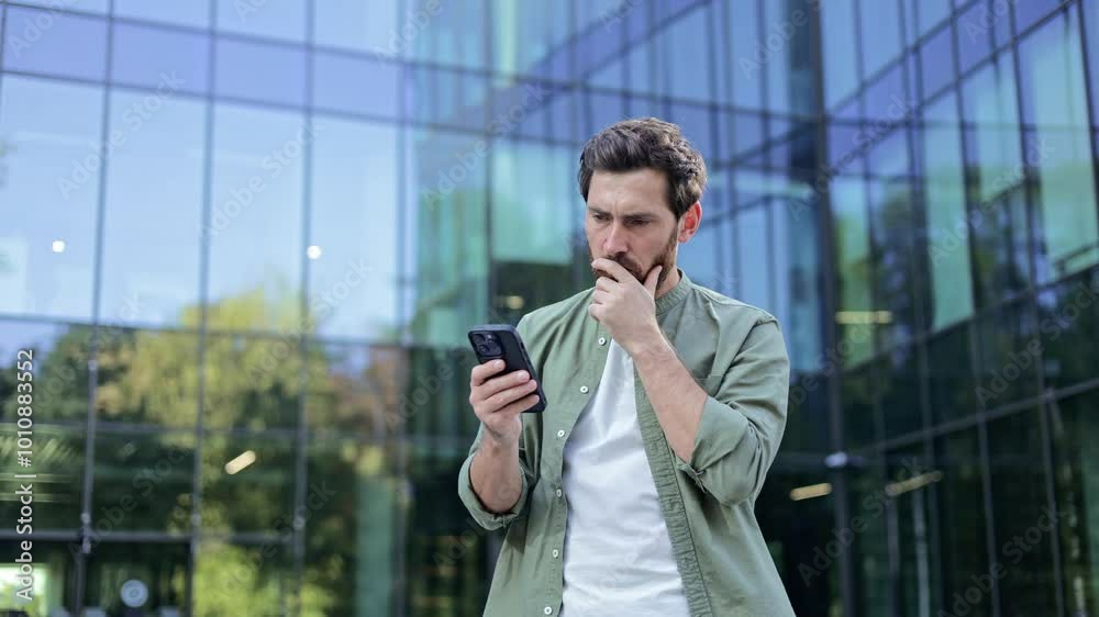 A young businessman has received concerning news on his smartphone. He is standing outside of an office building, and visibly showing his frustration. A young businessman has received concerning news on his smartphone. He is standing outside of an office building, and visibly showing his frustration.