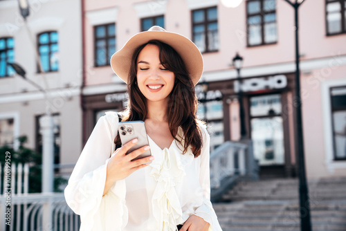 Young beautiful smiling woman in trendy summer clothes. Carefree female posing in street in sunny day. Positive model holds smartphone, looks at cellphone screen, uses mobile apps, in hat