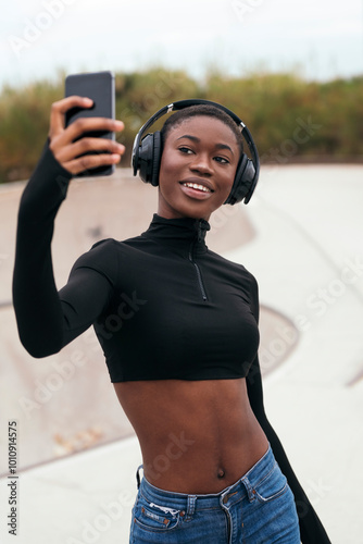 young ethnic female in casual apparel with wireless headset taking self portrait on mobile phone in a park photo