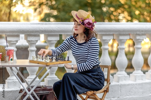 A young woman fashionably dressed in a vintage straw hat plays chess in an autumn park. photo