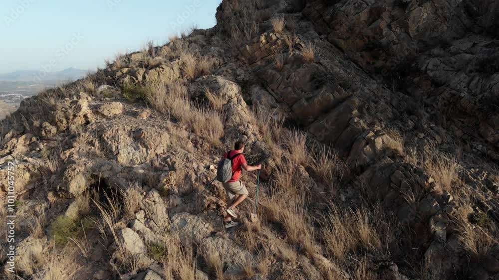 Climbing steep rocks off the trail, a man in a red shirt and shorts with trekking poles pursues his goal in the morning sun. Climbing steep rocks off the trail, a man in a red shirt and shorts with trekking poles pursues his goal in the morning sun.