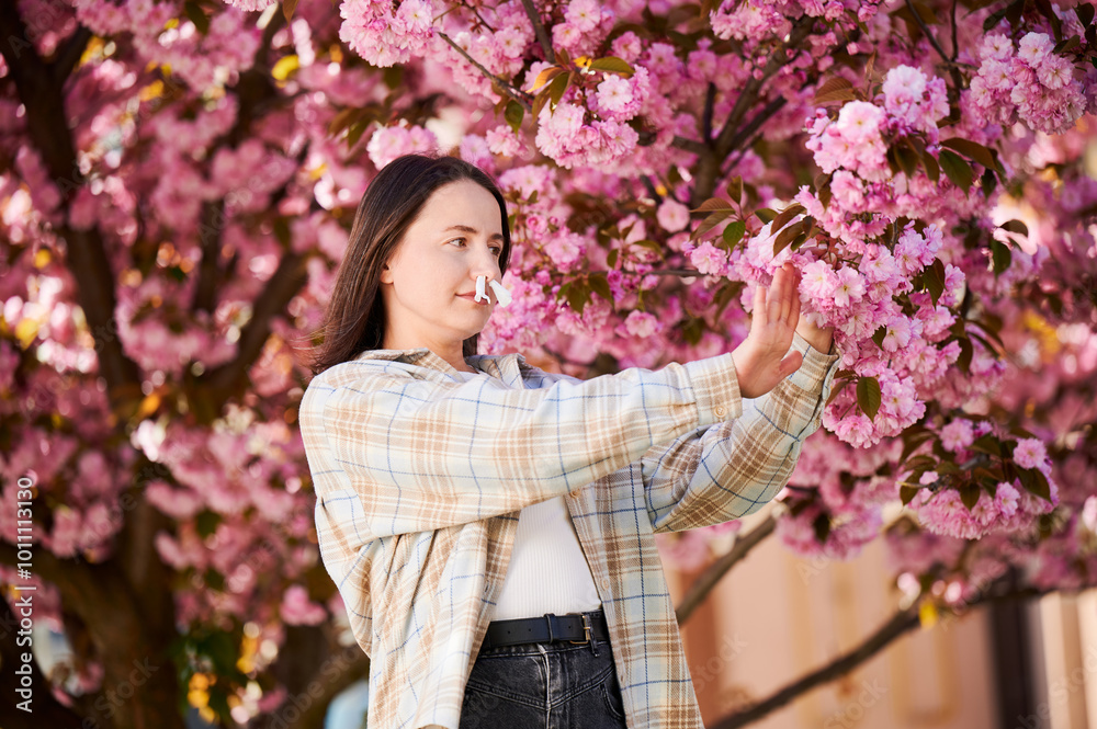 Fototapeta premium Woman allergic suffering from seasonal allergy and runny nose disease at spring. Young woman with paper tissues inserted into nose - symbolic gesture of inability to breathe due to nasal congestion.