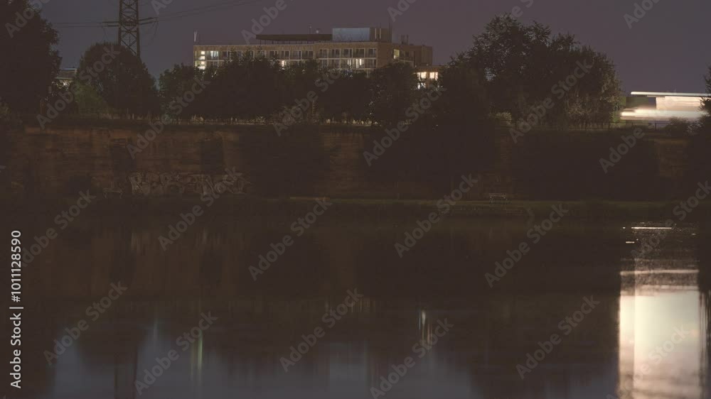 A city scene at night captures a passing train’s motion blur with reflections on the water’s surface. A long exposure video.