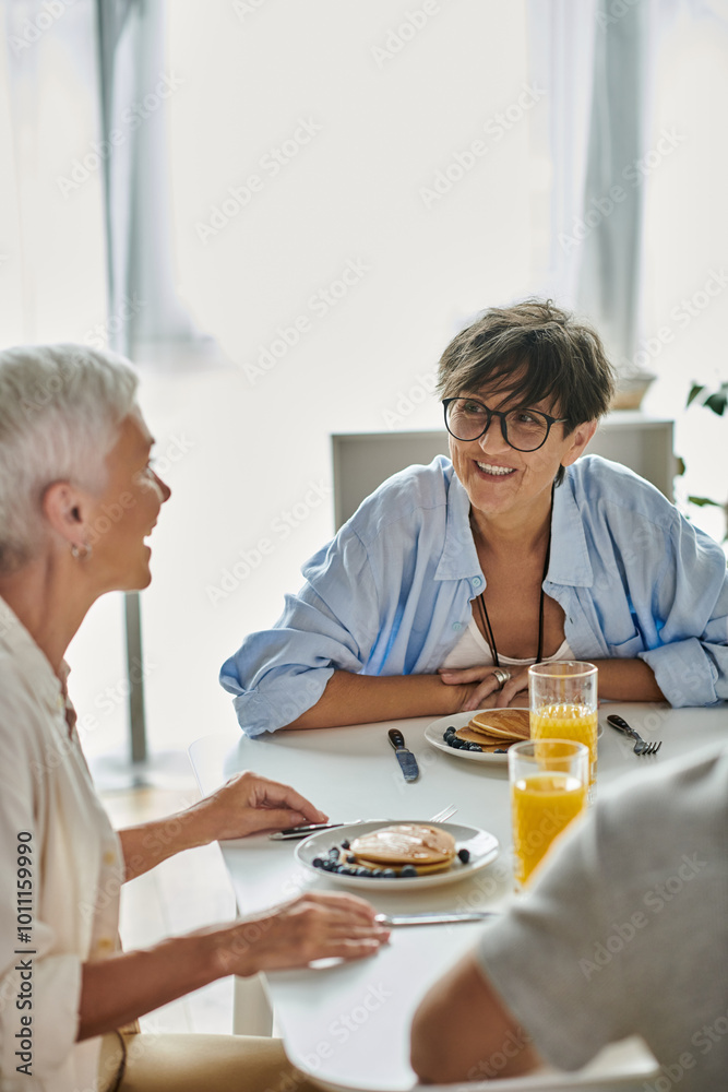 Lesbian parents enjoy a cheerful breakfast with their adult son, sharing laughter and love in their bright kitchen. Lesbian parents enjoy a cheerful breakfast with their adult son, sharing laughter and love in their bright kitchen.