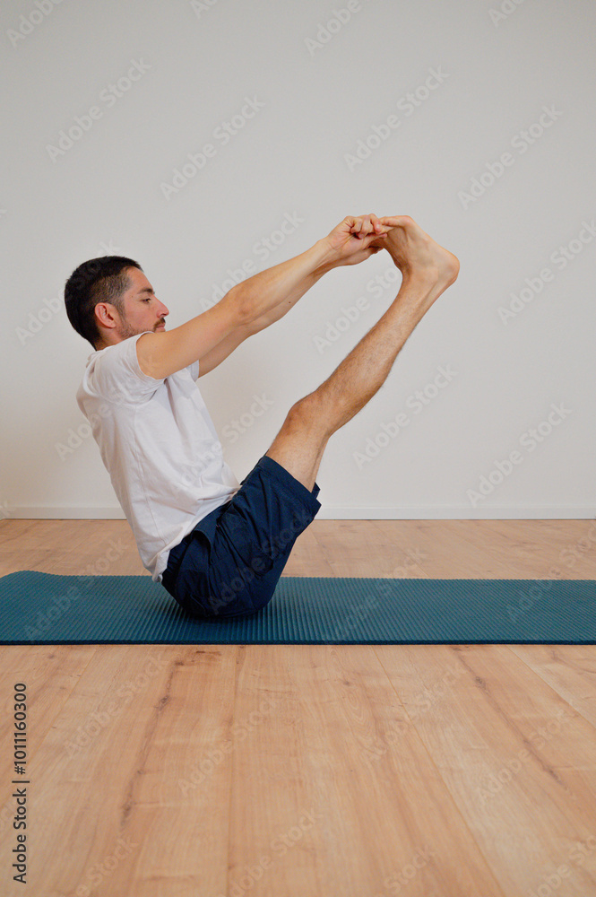 Fototapeta premium Focused Latin athlete performs morning exercise at home, holding the boat pose on a yoga mat. He trains his core with a static pose, highlighting abdominal strength and balance