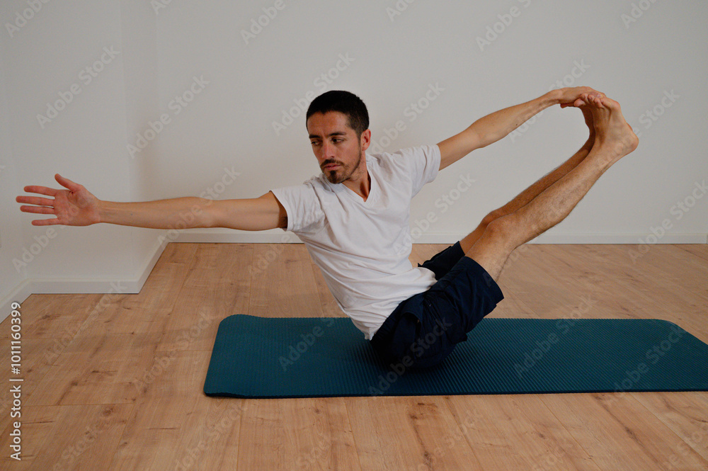 Athletic Latin man performs an advanced boat pose variation at home on a yoga mat. He balances while strengthening his core and flexing abs, showcasing concentration and body control Athletic Latin man performs an advanced boat pose variation at home on a yoga mat. He balances while strengthening his core and flexing abs, showcasing concentration and body control
