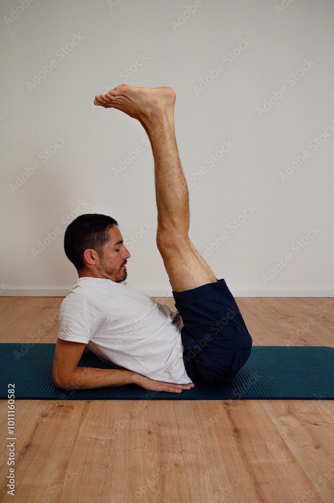 Fototapeta premium Athletic Latin man performs an advanced boat pose variation at home on a yoga mat. He balances while strengthening his core and flexing abs, showcasing concentration and body control