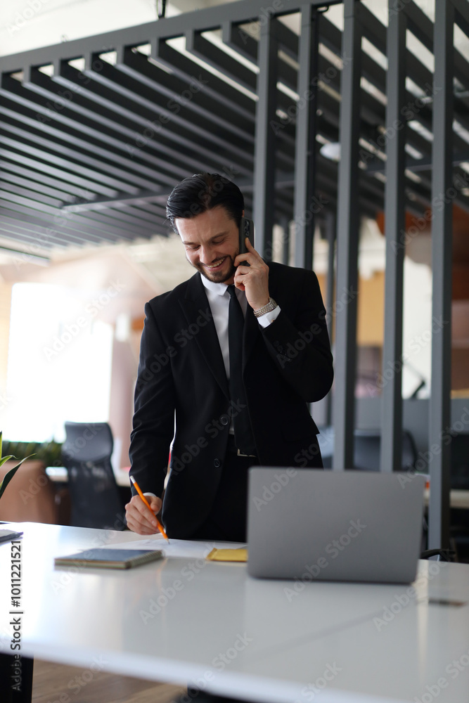 A man in a suit is talking on his cell phone while writing on a piece of paper. He is in a professional setting, possibly at work A man in a suit is talking on his cell phone while writing on a piece of paper. He is in a professional setting, possibly at work