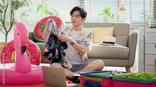 Young Asian man holding a camera and preparing for travel in a cozy living room, with a sofa, rubber ring, luggage, and sunglasses, summer time concept.	
