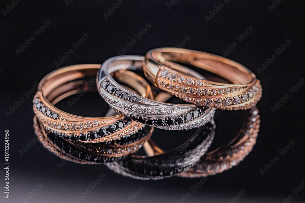 Wedding rings are reflected in the mirror. A set of three rings on a black background. Gold and silver rings with black and white stones. Wedding rings are reflected in the mirror. A set of three rings on a black background. Gold and silver rings with black and white stones.