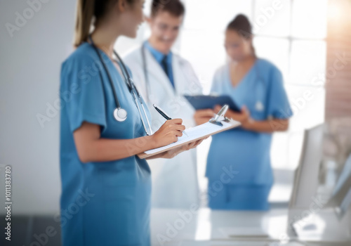 A female nurse in a blue scrub top and stethoscope writes on a clipboard with a pen. Two other medical personnel are out of focus behind her. photo