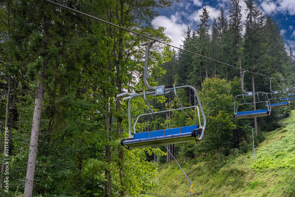 An empty lift descends to a sunny mountain resort in summer against the background of a pine forest. Cableway downhill, chair lift with sky in the background An empty lift descends to a sunny mountain resort in summer against the background of a pine forest. Cableway downhill, chair lift with sky in the background