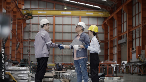 Three people are shaking hands in a factory. The man on the left is wearing a white shirt and blue jeans. The man on the right is wearing a white shirt and black pants