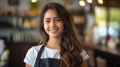 portrait of an Indian female hairdresser, with a smile and happy expression, wearing a black apron over a white t-shirt, with long brown wavy hair, standing in a salon background