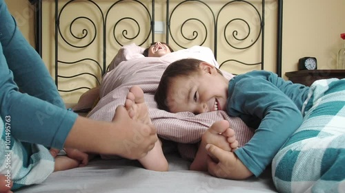 A child lying on a bed rolled up in a duvet is tickled on the soles of his feet by his younger siblings.