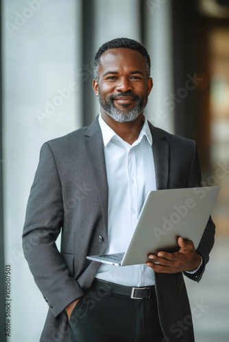 Middle-aged African American sales agent in formal attire, slightly smiling while holding a laptop, modern technological background.