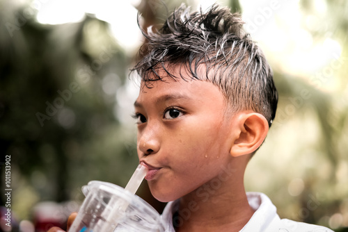 Thirsty and sweating Asian boy drinking iced beverage from a plastic glass using straw on a hot sunny day after playing