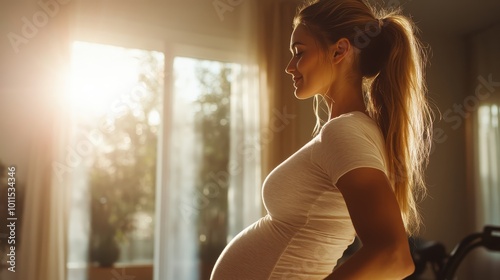 A pregnant woman stands in a sunlit room, displaying her profile while gently holding her belly, basking in the warm sunlight streaming through the window curtains. photo