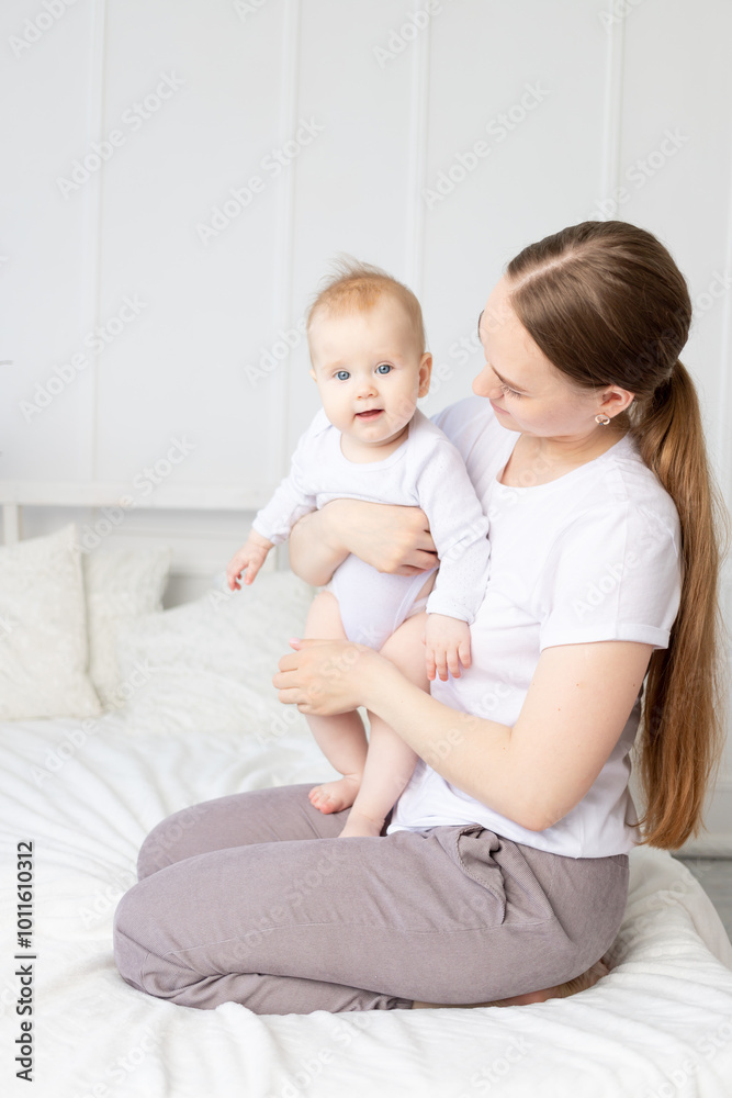 mom holds baby in her arms gently hugging him on the white bed at home, mother's love mom holds baby in her arms gently hugging him on the white bed at home, mother's love