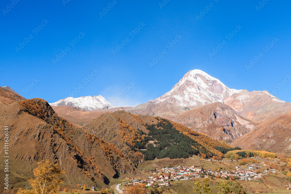 a panoramic view of autumn landscape with mountain hills surrounded by trees with yellow and red foliage at kazbegi stepantsminda in georgia near the foot of mount kazbek