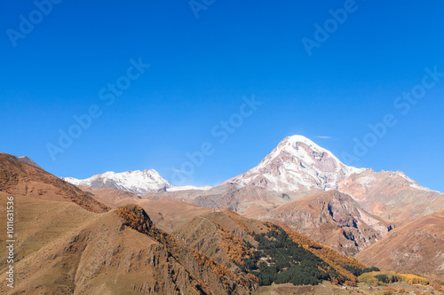 a panoramic view of autumn landscape with mountain hills surrounded by trees with yellow and red foliage at kazbegi stepantsminda in georgia near the foot of mount kazbek