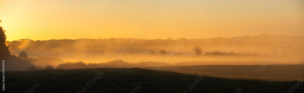 Fototapeta premium Sonnenaufgang Panorama: Fluss im Nebel mit kleinen Hügeln im Vordergrund