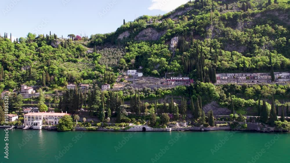 Breathtaking view of a serene lake surrounded by lush green mountains and a small Italian village on the shore. A bright summer day with fluffy clouds in the blue sky Breathtaking view of a serene lake surrounded by lush green mountains and a small Italian village on the shore. A bright summer day with fluffy clouds in the blue sky