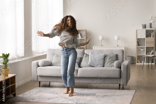 Cuadro en lienzo Cheerful carefree young Latin woman dancing in new modern apartment interior, celebrating home buying, property rent, hopping at pale cozy sofa, laughing, enjoying music
