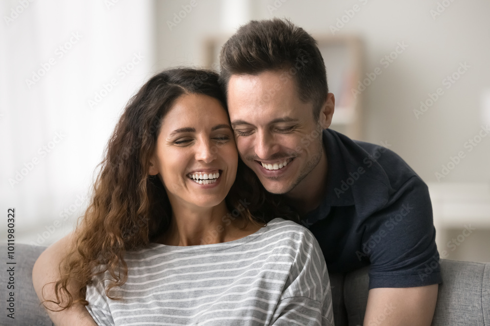 Joyful positive married couple having fun at home, enjoying leisure. Cheerful handsome husband hugging wife from behind sitting at comfortable couch with closed eyes, laughing Joyful positive married couple having fun at home, enjoying leisure. Cheerful handsome husband hugging wife from behind sitting at comfortable couch with closed eyes, laughing