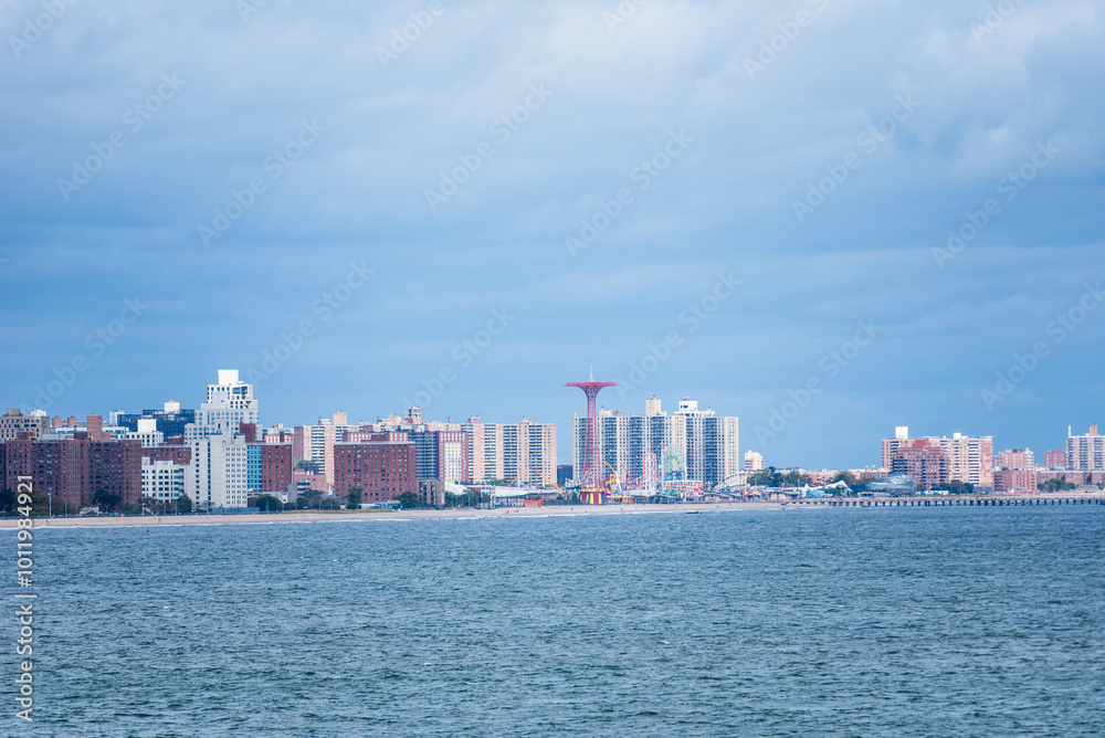 The skyline of Brooklyn emerges from the horizon, seen from a ship sailing through Gravesend Bay. The buildings stand in contrast against the cloudy sky and the steel-colored waters of the bay. The skyline of Brooklyn emerges from the horizon, seen from a ship sailing through Gravesend Bay. The buildings stand in contrast against the cloudy sky and the steel-colored waters of the bay.
