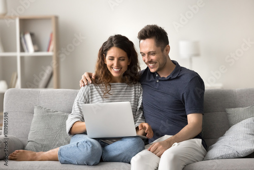 Cheerful attractive romantic couple using computer at home, sitting close on sofa, using modern Internet technology, online app, media service for communication, having fun, laughing