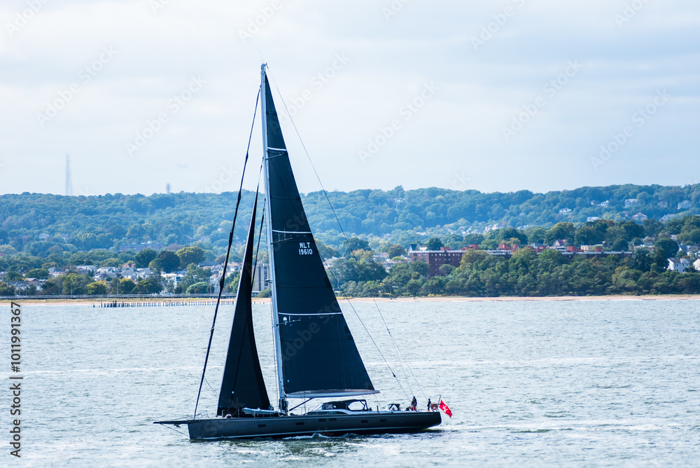 Naklejka premium A tourist yacht sails under full sails across the calm Atlantic waters near the coast of the USA, close to New York. The water shimmers silver, reflecting sunlight piercing through thick clouds.