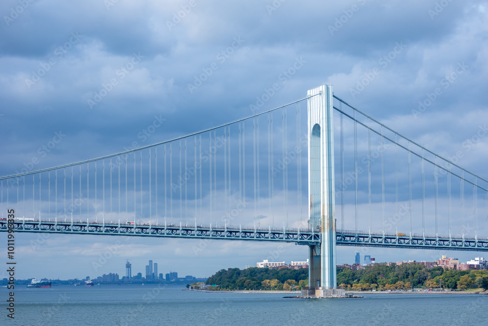 The Verrazzano-Narrows Bridge stretches across the Upper Bay near New York City, its steel arches silhouetted against a cloudy sky. The towering structure stands as a monumental gateway to the city.