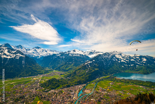 Breathtaking panorama of Interlaken, Switzerland, nestled between snow-capped Alps and lakes. Lush valleys, urban areas, and a paraglider in flight create a stunning landscape view