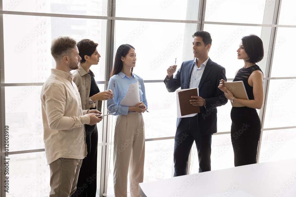 Indian business leader talking to diverse managers in meeting room, explaining teamwork strategy. Project team discussing work plan, standing and talking at large office window Indian business leader talking to diverse managers in meeting room, explaining teamwork strategy. Project team discussing work plan, standing and talking at large office window
