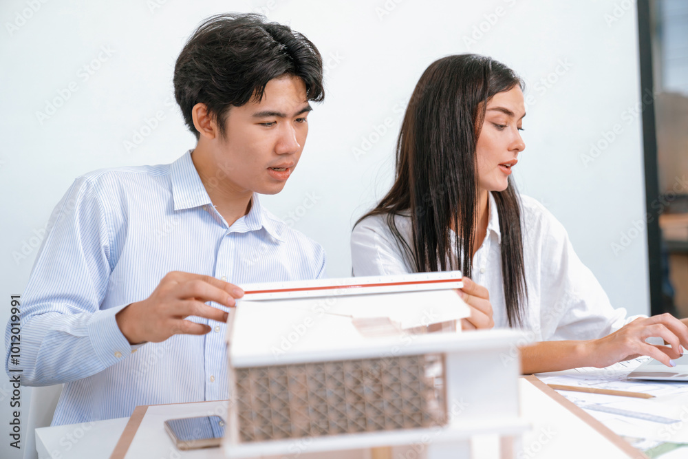 Professional male asian architect using ruler to measure house model length while young beautiful caucasian colleague using laptop to analyzed data on meeting table with house model. Immaculate. Professional male asian architect using ruler to measure house model length while young beautiful caucasian colleague using laptop to analyzed data on meeting table with house model. Immaculate.